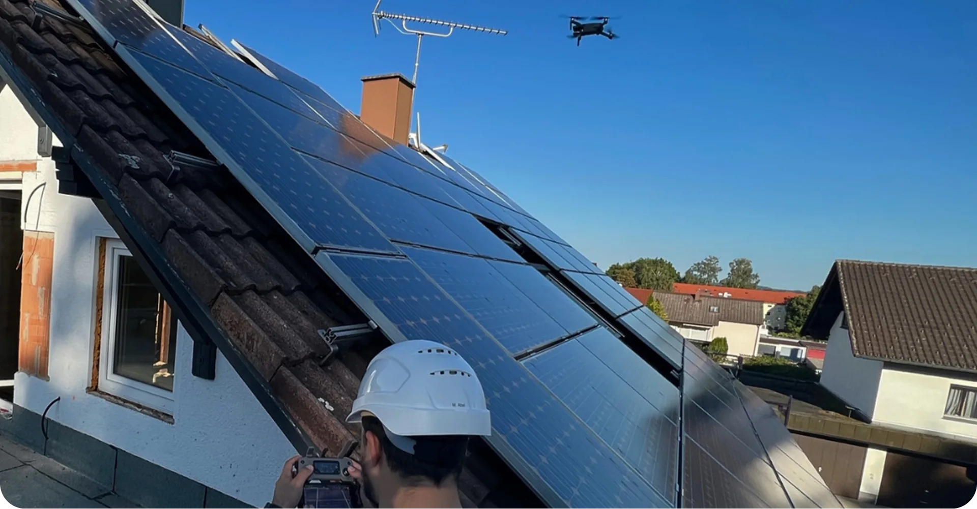 Person mit Helm steuert Drohne vor einem Haus mit Solarpaneelen auf dem Dach für die Erstellung einer Thermografie einer Solaranlage