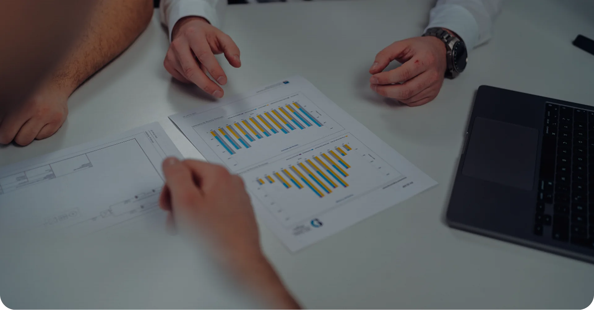 Drei Personen analysieren Papiere mit bunten Balkendiagrammen auf einem Tisch neben einem Laptop, während sie ein Wertgutachten besprechen.